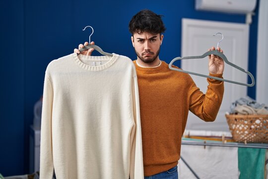 Hispanic man with beard holding sweater on hanger at laundry room depressed and worry for distress, crying angry and afraid. sad expression.