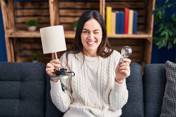 Young brunette woman holding led lightbulb and lamp winking looking at the camera with sexy...