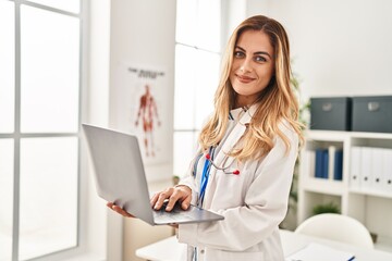 Young blonde woman wearing doctor uniform using laptop at clinic