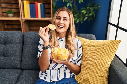Young blonde woman eating chips potatoes sitting on sofa at home