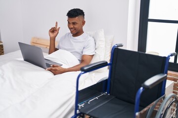 Young hispanic man lying on the bed, using wheelchair smiling happy pointing with hand and finger to the side