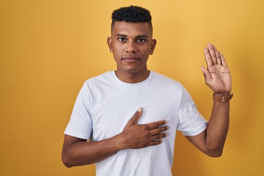 Young hispanic man standing over yellow background swearing with hand on chest and open palm, making a loyalty promise oath