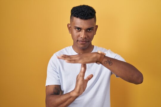 Young hispanic man standing over yellow background doing time out gesture with hands, frustrated and serious face