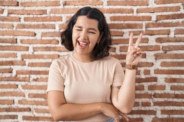 Young hispanic woman standing over bricks wall smiling with happy face winking at the camera doing victory sign with fingers. number two.