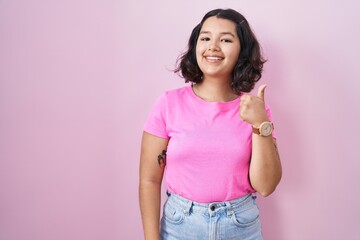 Young hispanic woman standing over pink background doing happy thumbs up gesture with hand. approving expression looking at the camera showing success.