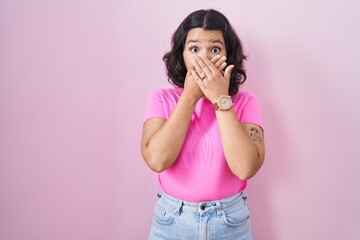 Young hispanic woman standing over pink background shocked covering mouth with hands for mistake....