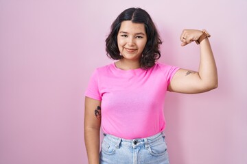 Young hispanic woman standing over pink background strong person showing arm muscle, confident and...