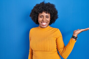 Fototapeta na wymiar Black woman with curly hair standing over blue background smiling cheerful presenting and pointing with palm of hand looking at the camera.