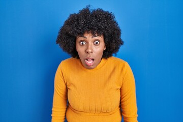 Fototapeta na wymiar Black woman with curly hair standing over blue background afraid and shocked with surprise and amazed expression, fear and excited face.