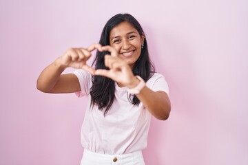 Young hispanic woman standing over pink background smiling in love doing heart symbol shape with hands. romantic concept.