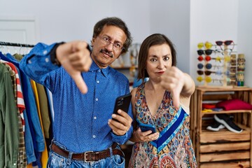 Obraz premium Middle age interracial couple at retail shop using smartphone looking unhappy and angry showing rejection and negative with thumbs down gesture. bad expression.