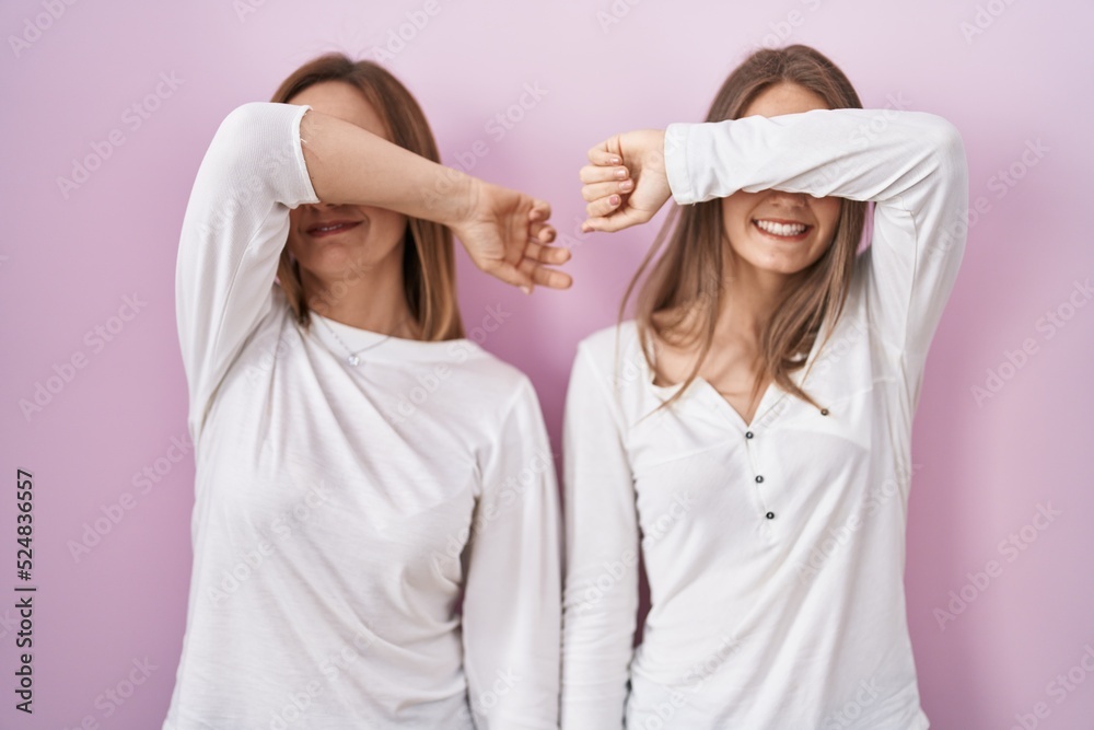 Wall mural Middle age mother and young daughter standing over pink background covering eyes with arm smiling cheerful and funny. blind concept.