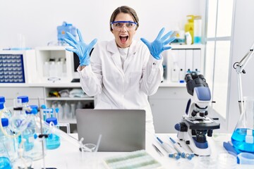 Young hispanic woman wearing scientist uniform working at laboratory celebrating victory with happy...