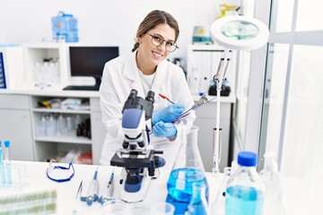 Fototapeta premium Young hispanic woman wearing scientist uniform using microscope at laboratory