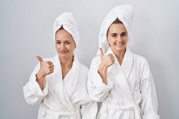 Middle age woman and daughter wearing white bathrobe and towel doing happy thumbs up gesture with...