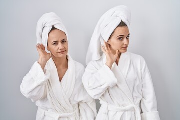 Middle age woman and daughter wearing white bathrobe and towel smiling with hand over ear listening an hearing to rumor or gossip. deafness concept.