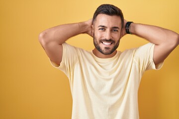 Fototapeta na wymiar Handsome hispanic man standing over yellow background relaxing and stretching, arms and hands behind head and neck smiling happy