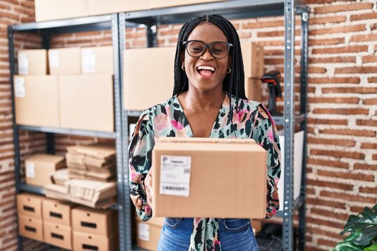 African woman with braids working at small business ecommerce holding package smiling and laughing hard out loud because funny crazy joke.