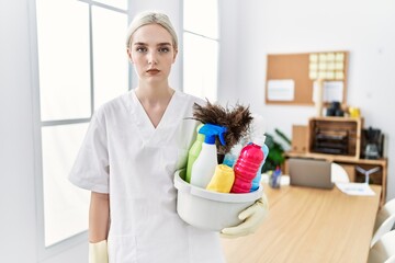 Young caucasian woman wearing cleaner uniform holding cleaning products cleaning office thinking...