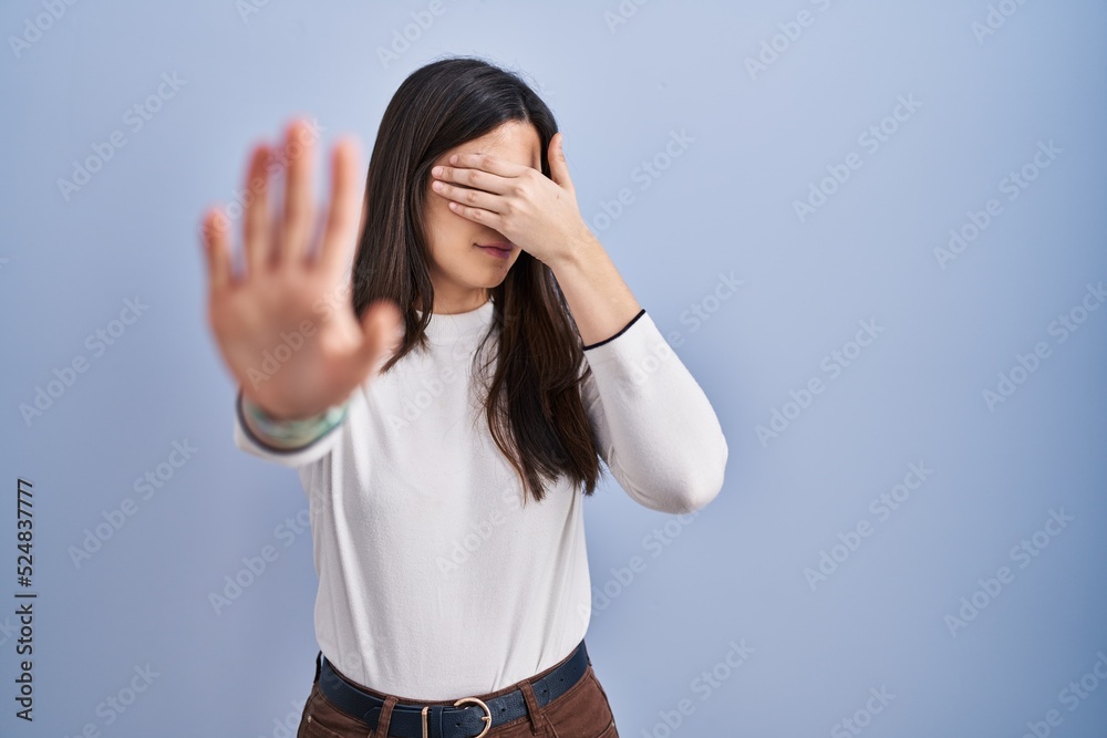 Poster young brunette woman standing over blue background covering eyes with hands and doing stop gesture w