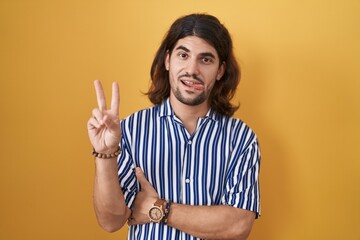 Hispanic man with long hair standing over yellow background smiling with happy face winking at the...