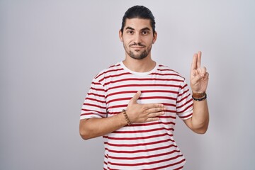 Hispanic man with long hair standing over isolated background smiling swearing with hand on chest...