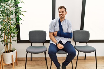 Handsome young man sitting at doctor waiting room with arm injury looking away to side with smile on face, natural expression. laughing confident.