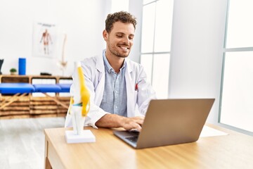 Young hispanic man wearing physiotherapist uniform using laptop working at clinic