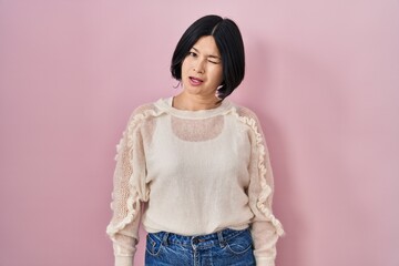 Young asian woman standing over pink background winking looking at the camera with sexy expression,...