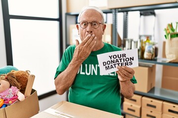 Senior volunteer man holding your donation matters covering mouth with hand, shocked and afraid for mistake. surprised expression