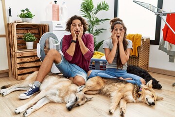 Young hispanic couple doing laundry with dogs afraid and shocked, surprise and amazed expression with hands on face