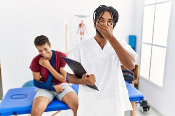 Young hispanic man working at pain recovery clinic with a man with broken arm laughing and...