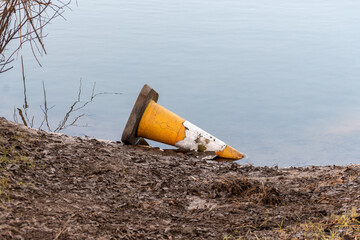 A tatty discarded traffic cone lying on its side in some shallow water next to a muddy bank.