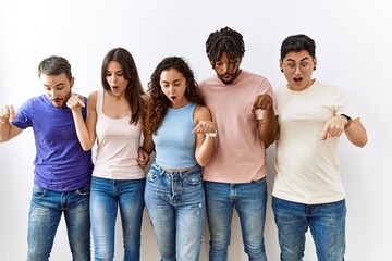 Obraz premium Group of young people standing together over isolated background pointing down with fingers showing advertisement, surprised face and open mouth