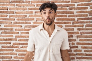 Arab man with beard standing over bricks wall background making fish face with lips, crazy and comical gesture. funny expression.