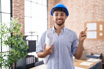 Arab man with beard wearing architect hardhat at construction office smiling happy pointing with...