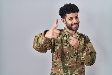 Arab man wearing camouflage army uniform pointing fingers to camera with happy and funny face. good energy and vibes.