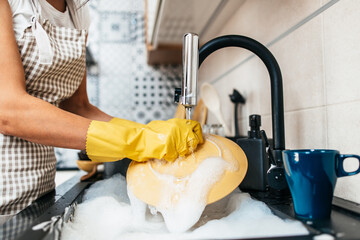 Young adult woman with yellow protective gloves washing her dishes on kitchen sink. Household and home hygiene routine. - obrazy, fototapety, plakaty
