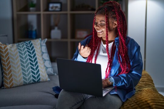African american woman with braided hair using computer laptop at night beckoning come here gesture with hand inviting welcoming happy and smiling