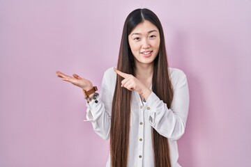 Chinese young woman standing over pink background amazed and smiling to the camera while presenting with hand and pointing with finger.