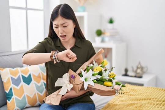 Chinese young woman holding bouquet of white flowers looking at the watch time worried, afraid of getting late