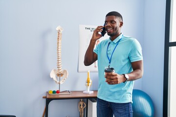 Young african american man physiotherapist talking on the smartphone and drinking coffee al rehab clinic - obrazy, fototapety, plakaty