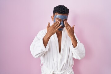 Young hispanic man wearing beauty face mask and bath robe rubbing eyes for fatigue and headache,...