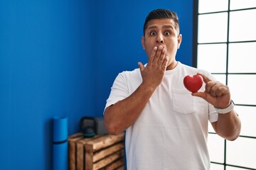 Hispanic young man holding red heart at gym covering mouth with hand, shocked and afraid for...