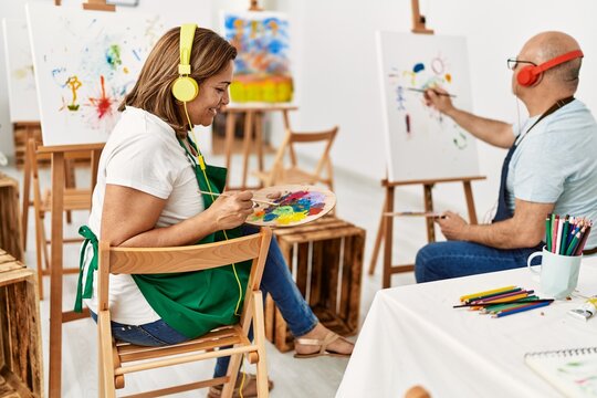 Middle age hispanic painter couple smiling happy using headphones and painting at art studio.