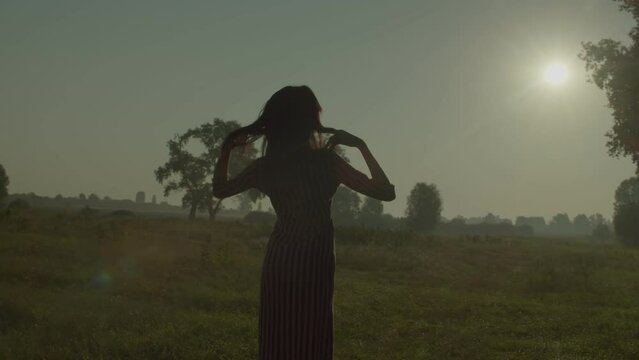 Rear view of elegant beautiful African American female in summer dress standing with arms outstretched on field, enjoying, freedom, leisure and serenity in scenic nature at daybreak.