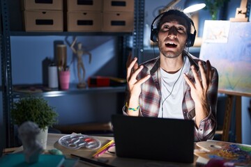 Young hispanic man sitting at art studio with laptop late at night crazy and mad shouting and yelling with aggressive expression and arms raised. frustration concept.