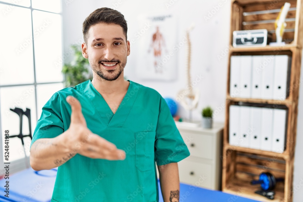 Wall mural Young physiotherapist man working at pain recovery clinic smiling friendly offering handshake as greeting and welcoming. successful business.