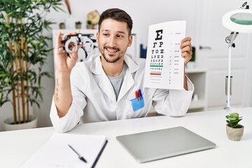 Young hispanic man wearing optician uniform holding snellen test at clinic