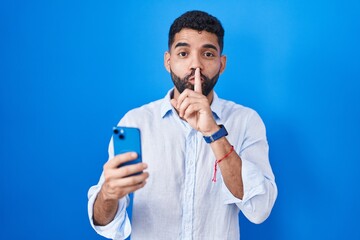 Hispanic man with beard using smartphone typing message asking to be quiet with finger on lips. silence and secret concept.